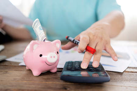 Man hand pen and calculator and piggy bank, money and magnifier on wooden table. putting money into piggy bank and counting on calculator closeupの写真素材