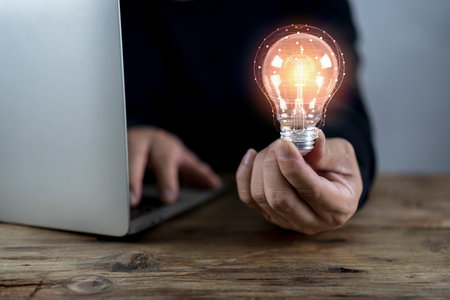 Businessman holding glowing lightbulb on wooden table with laptop computer as business solution and...