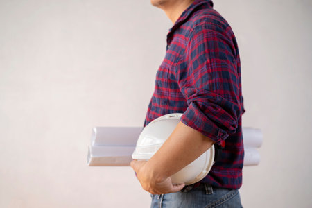 A young male engineer holding a blueprint with a white helmet while standing on a gray background....