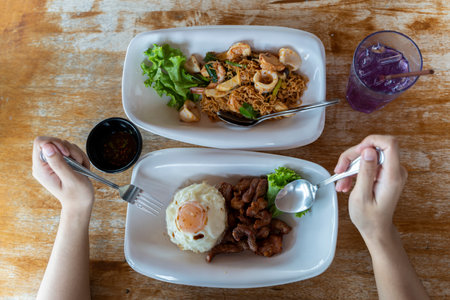 Hand holding a spoon The fork is placed on the dining table. Rice with fried pork, fried egg, mixed seafood saladの写真素材