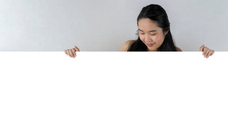 A cute beautiful woman holding a sign on a white background. Look down and look. Smiling woman holding a long blank white horizontal banner banner gray backgroundの写真素材