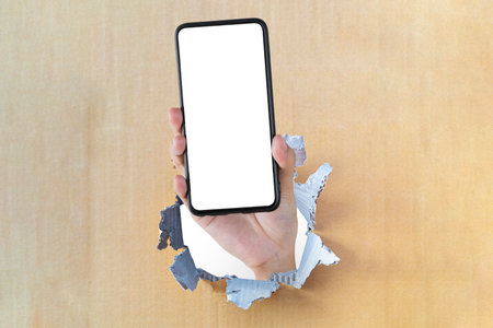 white hole in a brown cardboard background. To insert advertising media messages. holding smartphone with blank screen in hand through hole on brown cardboard background.の写真素材