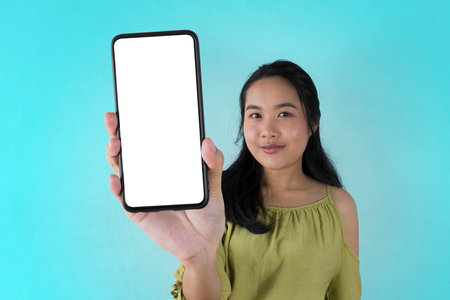 Trendy Mobile Phone. Excited cute Asian woman holding a smartphone with a blank white screen showing the device near the camera.の写真素材