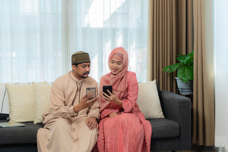 Asian muslim couple making a video call by using a cellphone while sitting together on the couch during Eid Mubarak in the living room. Shot at homeの写真素材