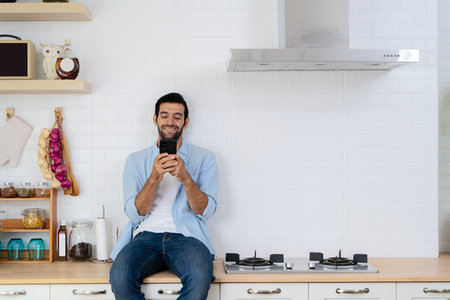 Handsome young man happy while sitting at home kitchen in good morning on leisurely vacation. Cheerful young bearded man talking on 

smart phone using social media checking work with smiling face.の写真素材