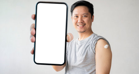 Man smiling after getting a vaccine. Man holding down him shirt sleeve and showing him arm with bandage after receiving accination, holding a large smartphone with a blank screen.の写真素材