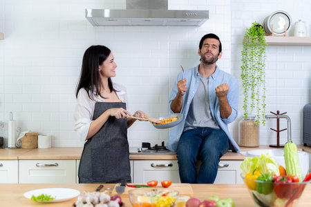 Romantic Couple At Kitchen. Man tasting food with good taste. Beautiful young wife cooking salad with smiling husband waiting for dinner or breakfast. Family enjoying weekend together.の写真素材