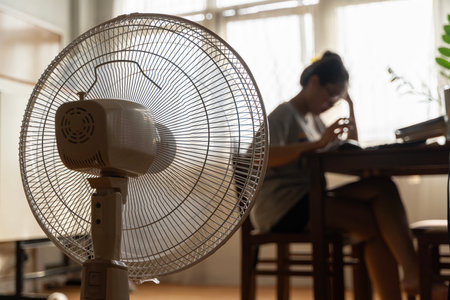 Asian unhappy woman sitting in front of working fan suffering from heat in modern house on sunny summer day in front of working fan suffering from summer heat.の写真素材