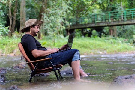 Handsome bearded Asian man of working age sitting on camping chair and working on laptop near stream in middle of park in outdoor forest alone happily.の写真素材
