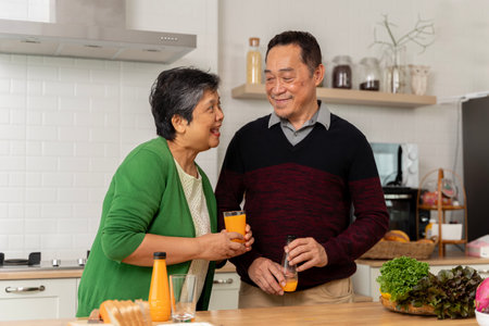 Loving senior couple in aprons toasting each other with orange juice and preparing healthy dinner while spending time at home. Different fruits on table. Healthy food at home.の写真素材