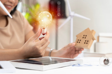Woman with light bulb and windmill. Cropped view of woman holding light bulb on white table with model of wind turbine, solar cell and block house on work table. Green energy concept.の写真素材