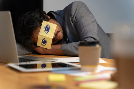 Cheerful adult man resting in office with sticky notes on the eyes. Sleeping man having stickers with eyes painted on face relaxing with hands behind head in office.の写真素材