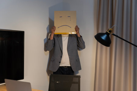 Portrait of a businessman covering his face with a paper mask with a sad face, drawn on it Bad negative depression from the anonymous person in the corner. Unhappy employee has a box instead of a headの写真素材