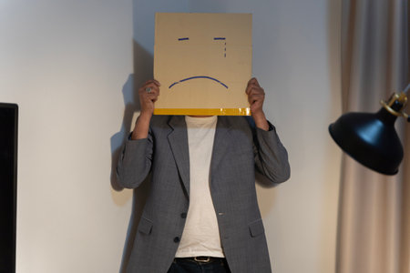 Unhappy man employee with box instead of his head. Portrait of businessman covering his face with paper mask with sad face drawn on it, bad negative sullen depression anonymous person.の写真素材