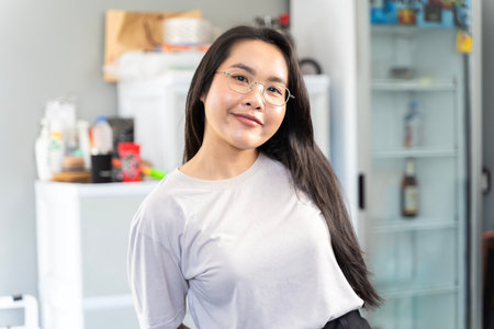 A beautiful young Asian woman standing inside a house. She is smiling brightly and wearing glasses. The woman may be working, or relaxing at home.の写真素材