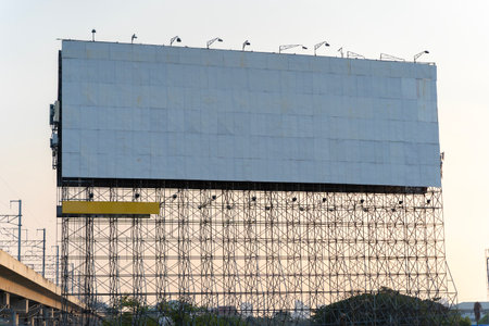 A large white billboard on scaffolding in front of a sunset sky on the side of a highway. The billboard is surrounded by trees and bushes. The sun is shining brightly.の写真素材