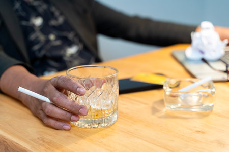 Close-up of a hand holding a glass of liquor. A disheveled man sits in his living room, surrounded by empty bottles of liquor. He is wearing a crumpled suit and tie, his expression one of despair.の写真素材
