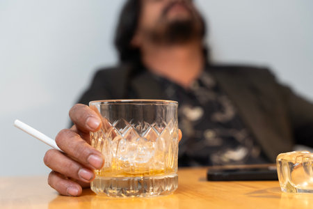 Close-up of a hand holding a glass of liquor. A disheveled man sits in his living room, surrounded by empty bottles of liquor. He is wearing a crumpled suit and tie, his expression one of despair.の写真素材