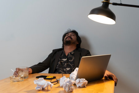 Asian businessman fails, man in despair drinks hard liquor, sitting late at the table in the office, depressed and hopeless. concept of alcoholism in the workplace. Drunk man in the office.の写真素材