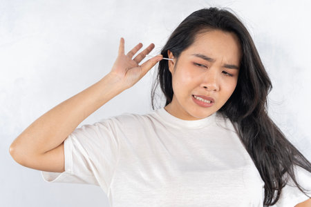 Close up young woman cleaning ears, using cotton bud after shower, feeling pain, beautiful female wearing white t-shirt standing in bathroom, morning routine, personal hygiene conceptの写真素材