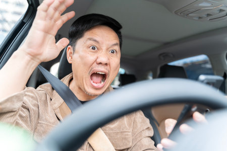 A man is yelling in a car is driving a car and looking at his cell phone. He is surprised and excited. Concept of distraction while driving and the potential danger it poses.の写真素材