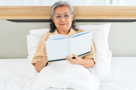 A woman is reading a book while sitting on a bed. She is wearing glasses and she is enjoying her book.の写真素材