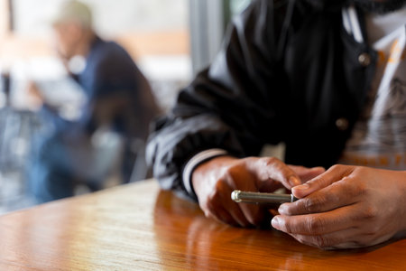 Closeup image of a man holding and using smart phone with coffee cup on wooden table in cafe. Man...