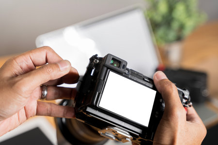 Photographer is holding professional digital camera near laptop with blank screen, preparing for photo editing. A camera with a white screen is being held by a person, clipping path.の写真素材
