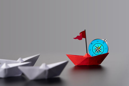 Red paper boat perched on a compass rose, surrounded by white boats, symbolizing direction and leadership. Red Paper Boat On Compass Leading A Fleet Of Small White Boats. Business Leadershipの写真素材