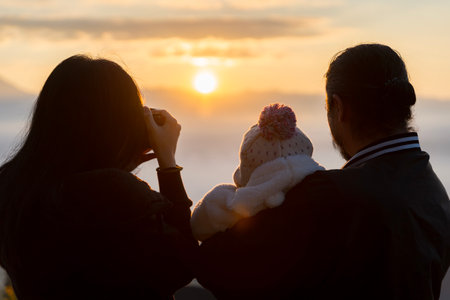 Father, mother and daughter near with his back to the camera on a hill and hugging each other enjoying a beautiful sunset. Woman photographing father with children. Silhouettes of Happy family.の写真素材