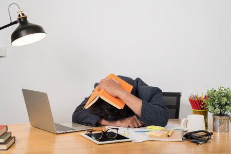 Working late in the office. Stressed young man sitting at a table among books. Overworked guy sleeping during his studies at city cafe, putting book on his head, tired of doing home assignment.の写真素材