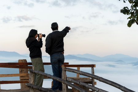 Man pointing at scenic mountain view while woman takes photo with DSLR camera on wooden terrace at sunrise. Travel, tourism, nature photography, adventure, and couple outdoor lifestyle concept.の写真素材