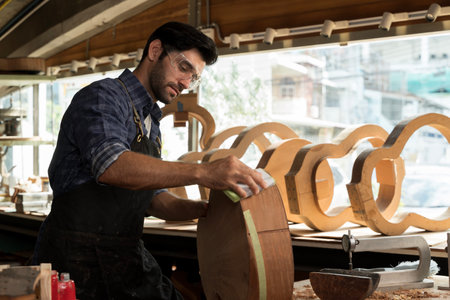 Focused male carpenter sanding a wooden piece in a woodworking shop. He wears protective goggles and apron, representing skilled labor, craftsmanship, handmade furniture, and professional woodwork.の写真素材