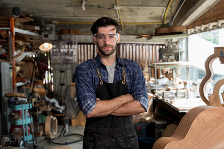 Portrait of confident male carpenter wearing safety goggles and apron standing with arms crossed in woodworking workshop. Concept of craftsmanship, skilled labor, and professional woodworkerの写真素材