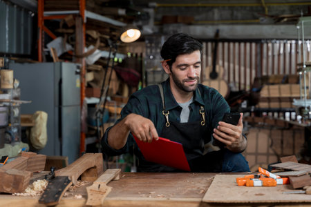 Young male carpenter using smartphone and holding clipboard in woodworking workshop. Concept of modern craftsmanship, multitasking, small business owner, digital tools, and work managementの写真素材
