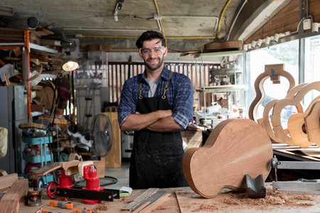 Smiling luthier standing with arms crossed in workshop, surrounded by guitar parts and tools. Concept of craftsmanship, woodworking, handmade instrument building, and creative studio workspaceの写真素材