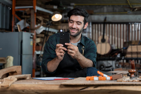 Happy carpenter sitting in woodworking workshop using smartphone. Concept of small business owner managing marketing, social media, communication, and orders online in modern digital economyの写真素材