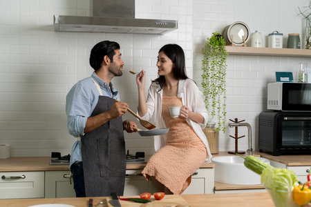 Smiling woman sits on kitchen counter and feeds man with spoon while he holds frying pan. Happy couple enjoying cooking and spending quality time together in cozy home kitchen environment.の写真素材