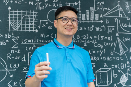 Portrait of a smiling Asian male teacher in blue shirt holding chalk in front of a chalkboard filled with mathematical formulas. Perfect for education, school, learning, and academic themes.の写真素材