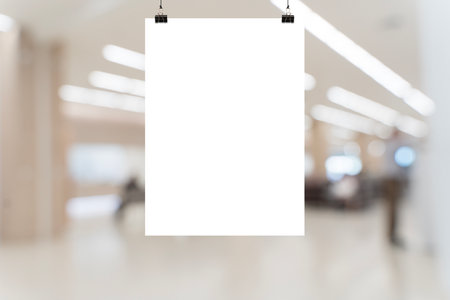 Empty white vertical poster mockup hanging in public indoor space with blurred background of people and lights. Suitable for signage design, advertisement, direction, or announcement template.の写真素材