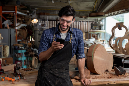 Happy carpenter taking a break in woodworking shop while checking smartphone. Wearing safety goggles and apron, standing near wooden instrument. Concept of craft, tech use, and modern labor.の写真素材