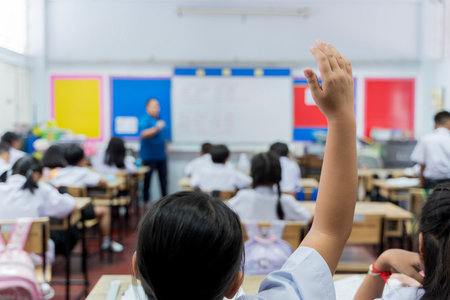 Elementary classroom scene with students seated at desks, one child raising hand to participate, while teacher explains at the front. Bright and engaging school environment focused on learning and interaction.の写真素材