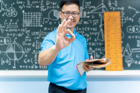 Confident smiling teacher in classroom showing OK hand gesture while holding book, standing in front of blackboard with math and science formulas, concept of education success knowledge and teachingの写真素材