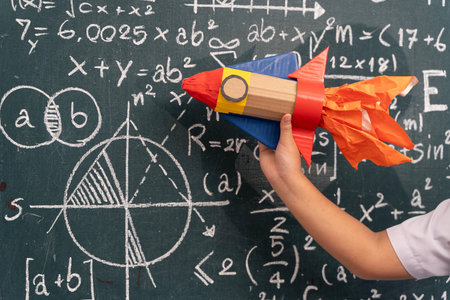 Close-up of student hand holding colorful handmade rocket craft against classroom blackboard filled with complex math formulas, equations, and geometric science drawings, creativity in educationの写真素材