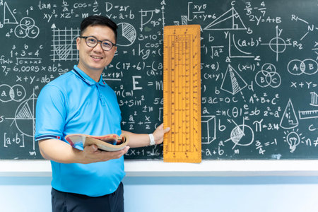 Asian male mathematics teacher in blue shirt holding a book and a large wooden ruler standing in front of blackboard with mathematical formulas, equations, and geometric shapes, smiling at cameraの写真素材