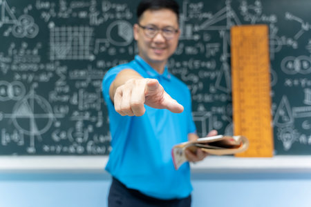 Asian male mathematics teacher in blue shirt smiling and pointing finger straight at camera while...
