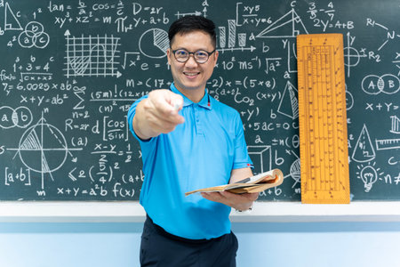 Asian male mathematics teacher in blue shirt smiling and pointing white chalk toward camera while holding book in front of blackboard with math formulas, equations, and geometric figures in classroomの写真素材