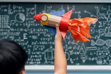Student raising colorful handmade rocket craft in classroom, standing in front of blackboard covered with scientific equations, mathematical formulas, and geometric drawings, creativity in educationの写真素材