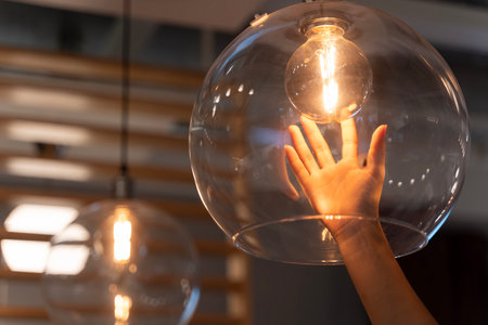 Close up of human hand gently touching glowing filament light bulb inside clear glass sphere pendant lamp in stylish modern interior. Concept of warmth, creativity, inspiration, and interior decor.の写真素材