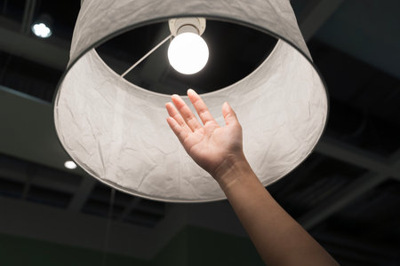 Close up of hand reaching toward glowing light bulb inside lamp shade. Concept of bright idea,...